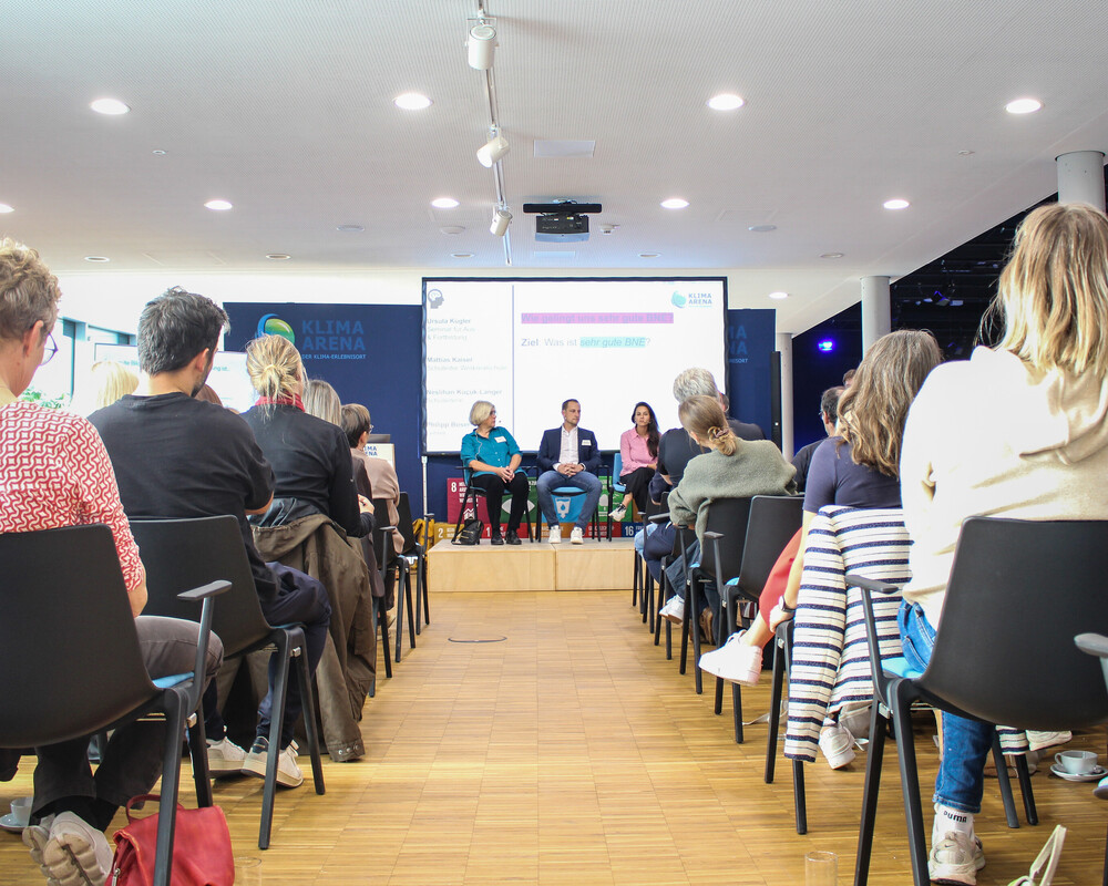 Blick durch die Stuhlreihen auf die G&auml;ste einer Podiumsdiskussion auf der B&uuml;hne